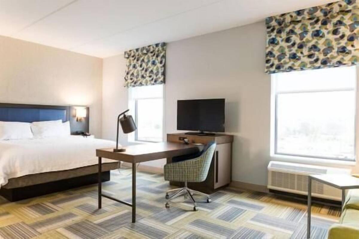 A hotel room with a bed, desk, chair, TV, and large windows.