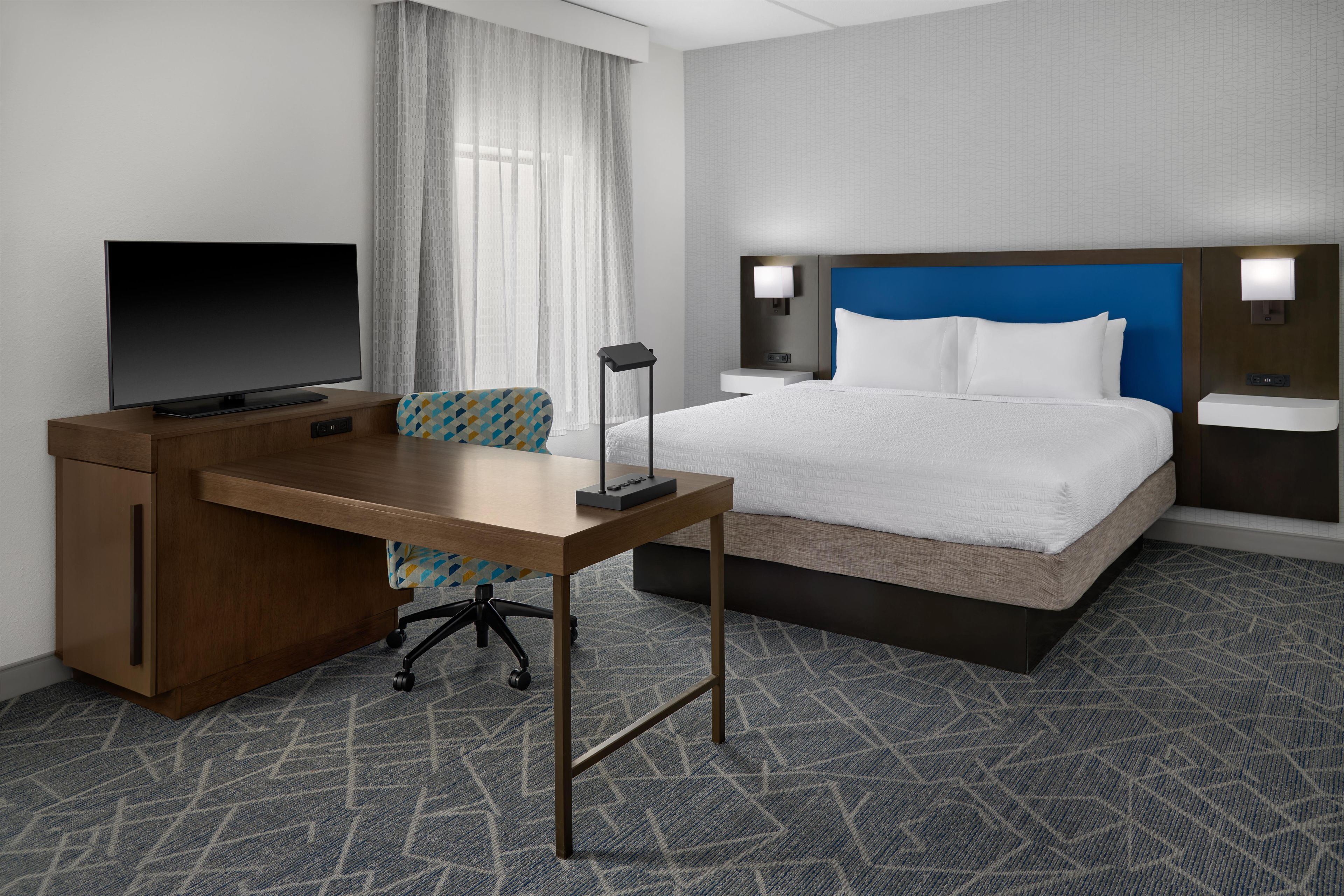Quarto de hotel com cama, escrivaninha, cadeira, televisão e janela com cortinas.