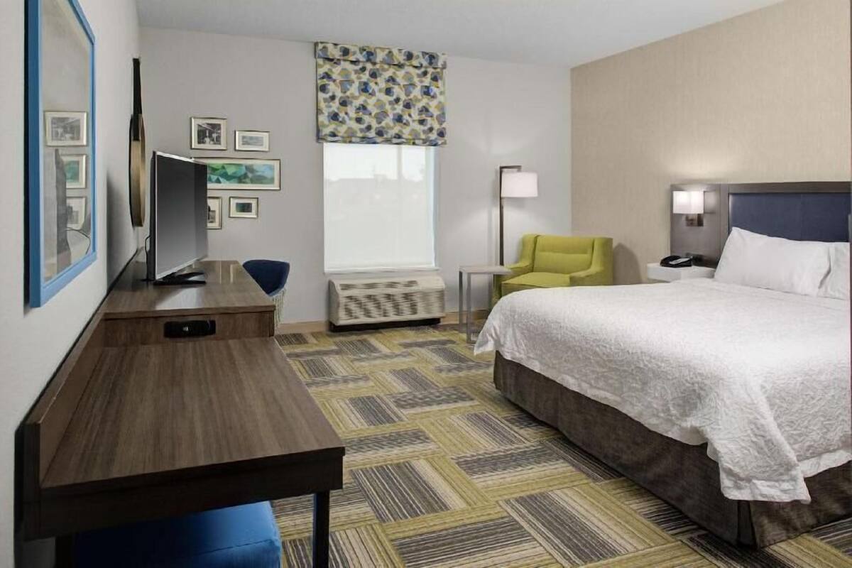 A hotel room with a bed, a desk, a chair, a lamp, and a window with a view.