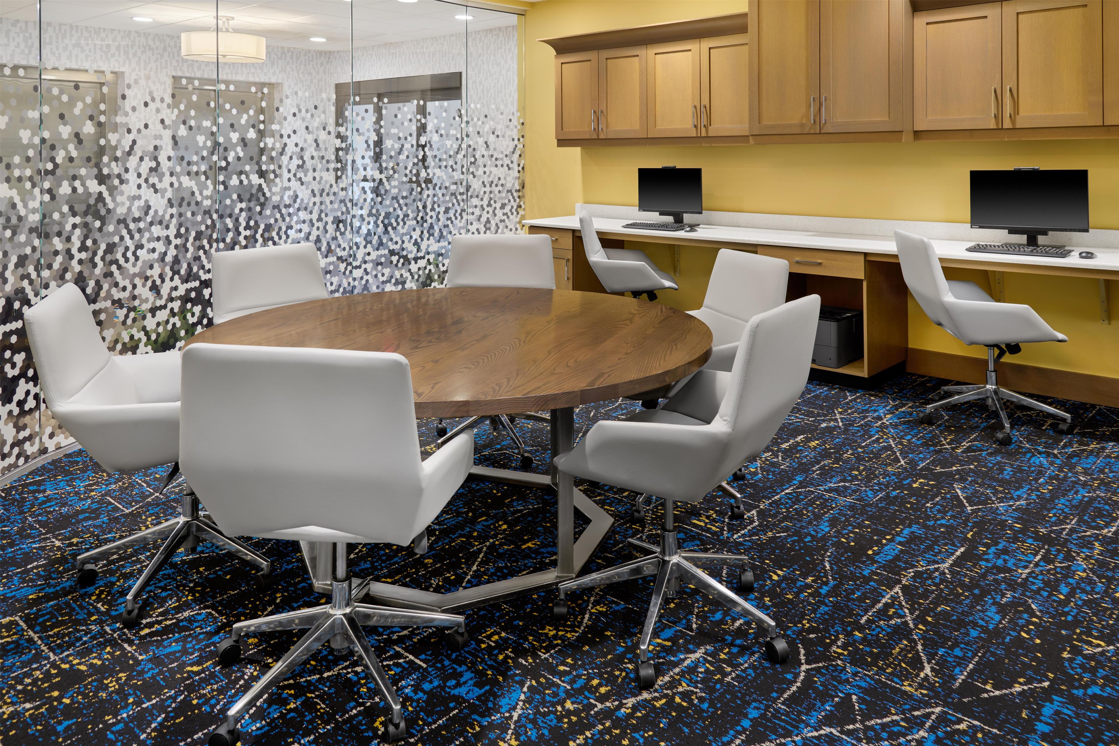 A conference room with a round table, six white chairs, a glass partition, and two computer desks with monitors.