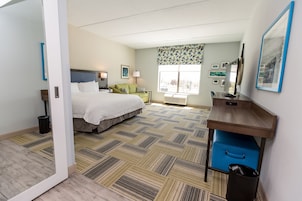 Quarto de hotel com cama, escrivaninha, cadeira, janela com vista e lixeira.