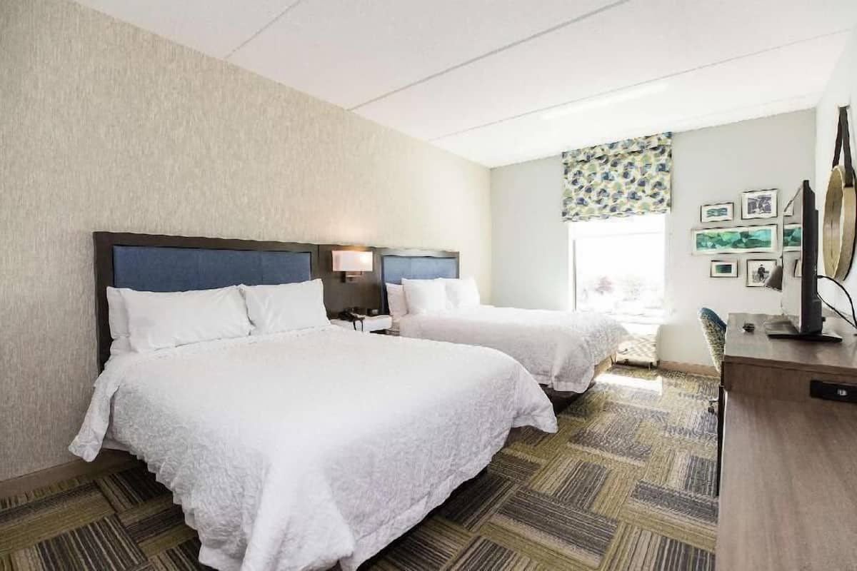 A hotel room with two beds, a desk, a lamp, and a window with a view.