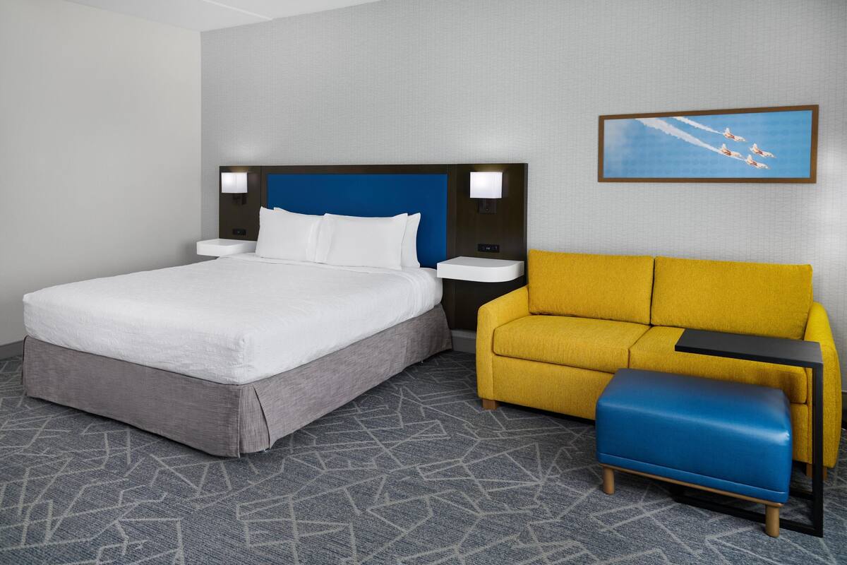 A hotel room with a large bed, a yellow sofa, a small blue ottoman, and a wall-mounted picture of airplanes.