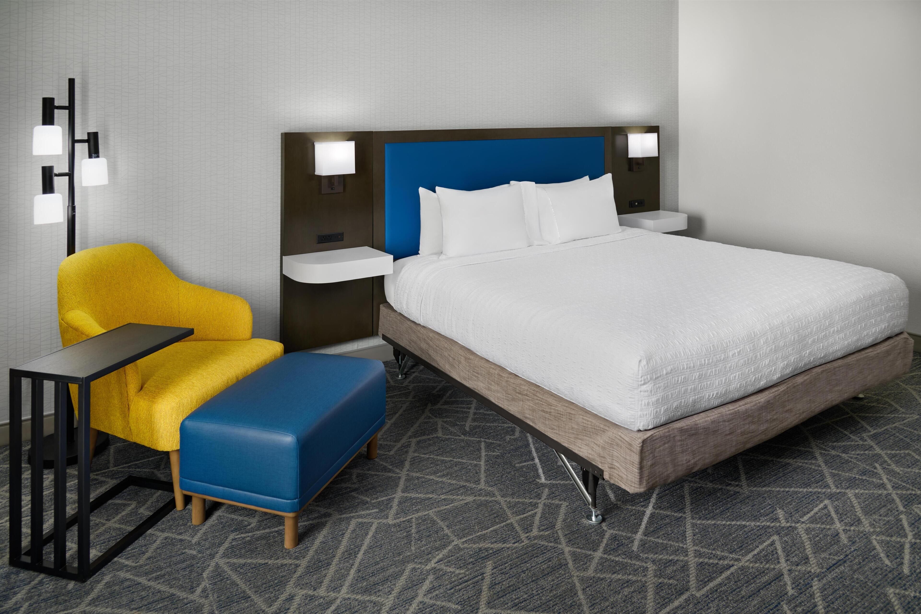 Quarto de hotel com uma cama grande, uma poltrona amarela, um banquinho azul, uma mesinha de cabeceira e uma luminária na parede.