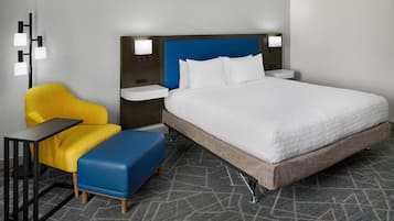 Quarto de hotel com uma cama grande, uma poltrona amarela, um banquinho azul, uma mesinha de cabeceira e uma luminária na parede.