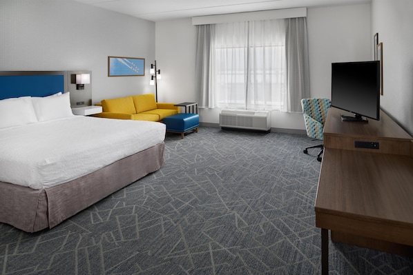 Habitación de hotel con cama, sofá, escritorio con televisor, lámpara y ventana con cortinas.