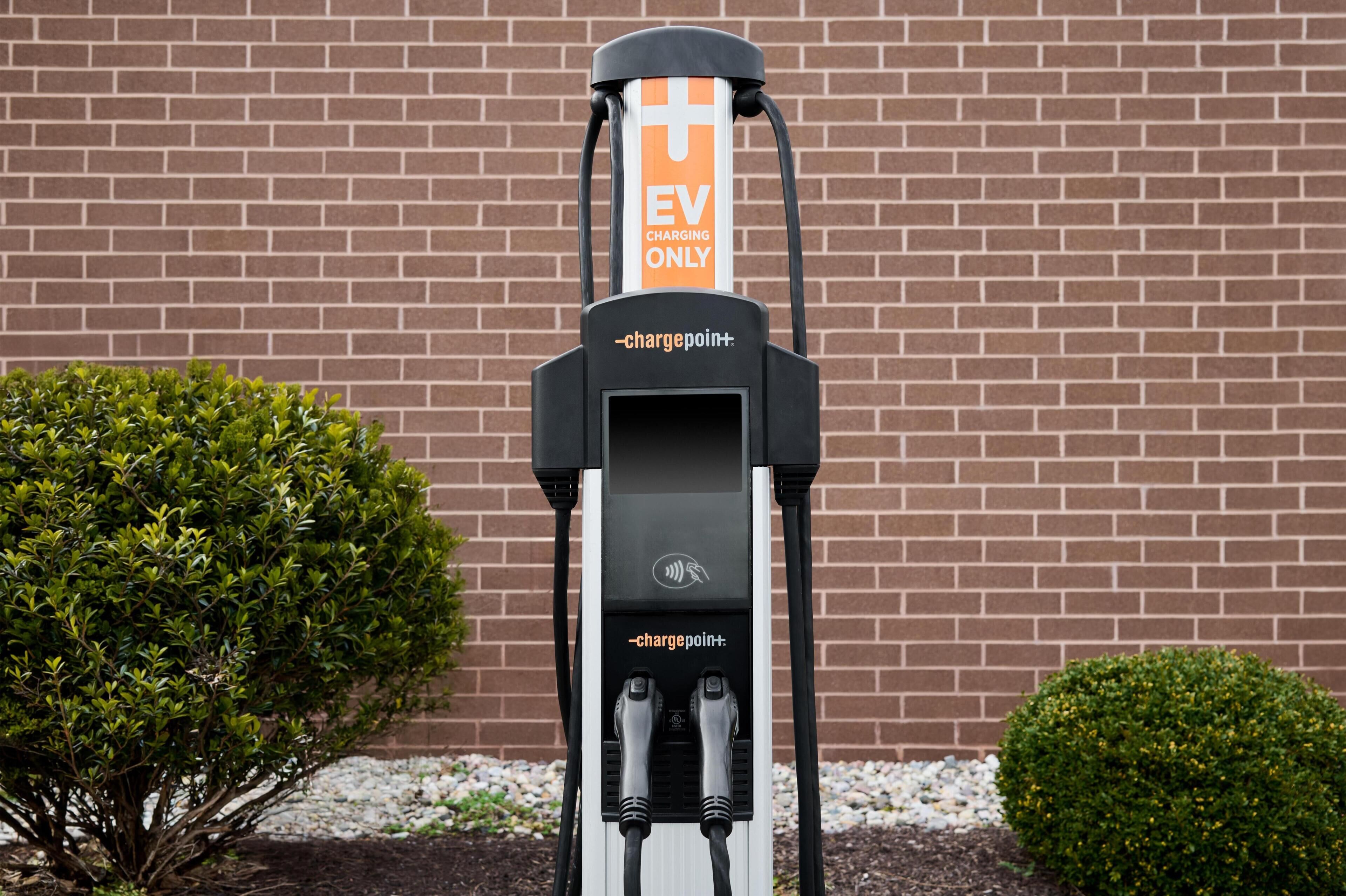 An EV charging station with a brick wall background and small bushes.