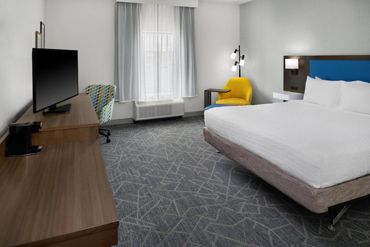 A hotel room with a bed, a desk with a flat-screen TV, a chair, a nightstand with a lamp, and a window with curtains.