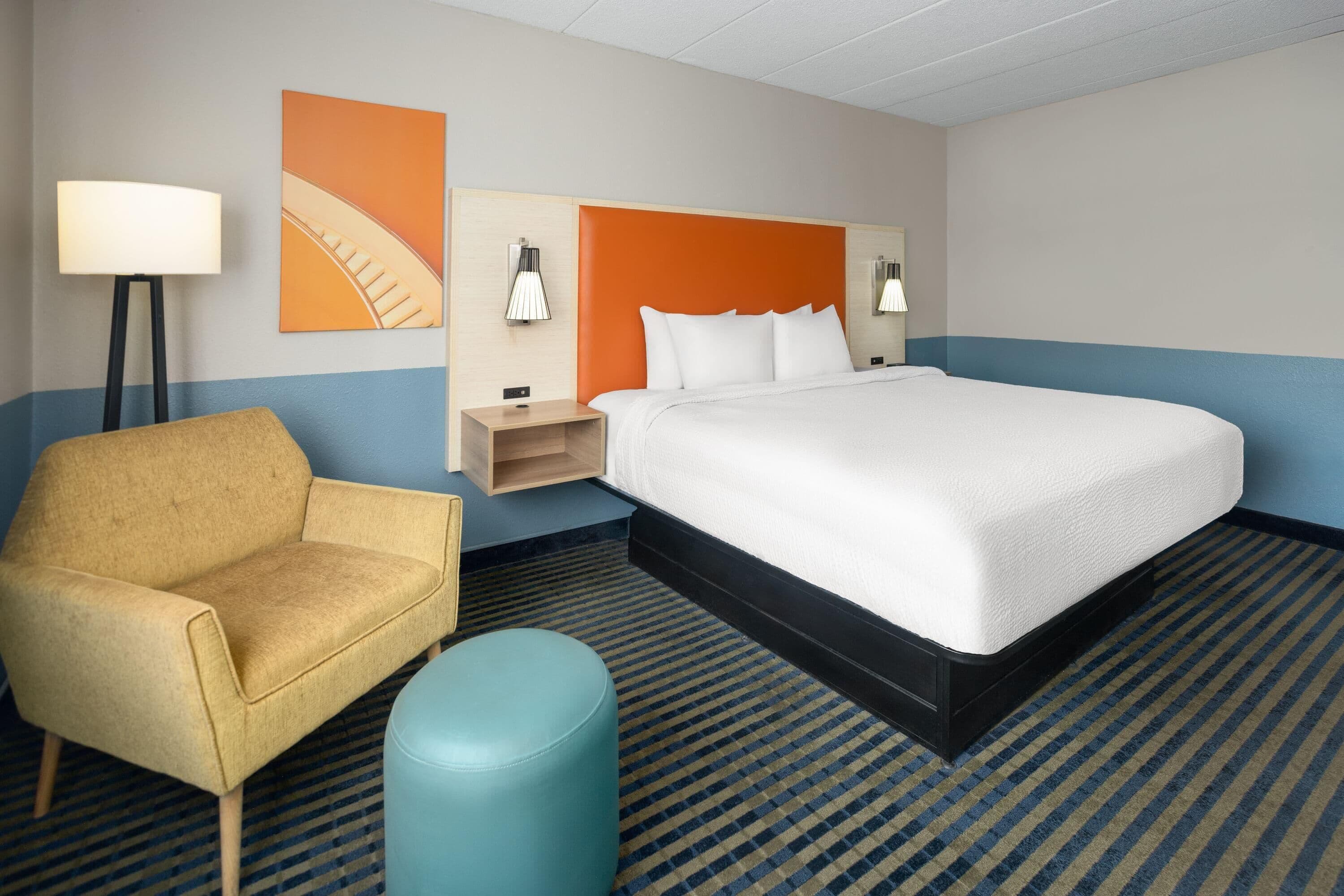 Una habitación de hotel con una cama grande, una silla, un pequeño reposapiés y una mesita de noche con lámparas.
