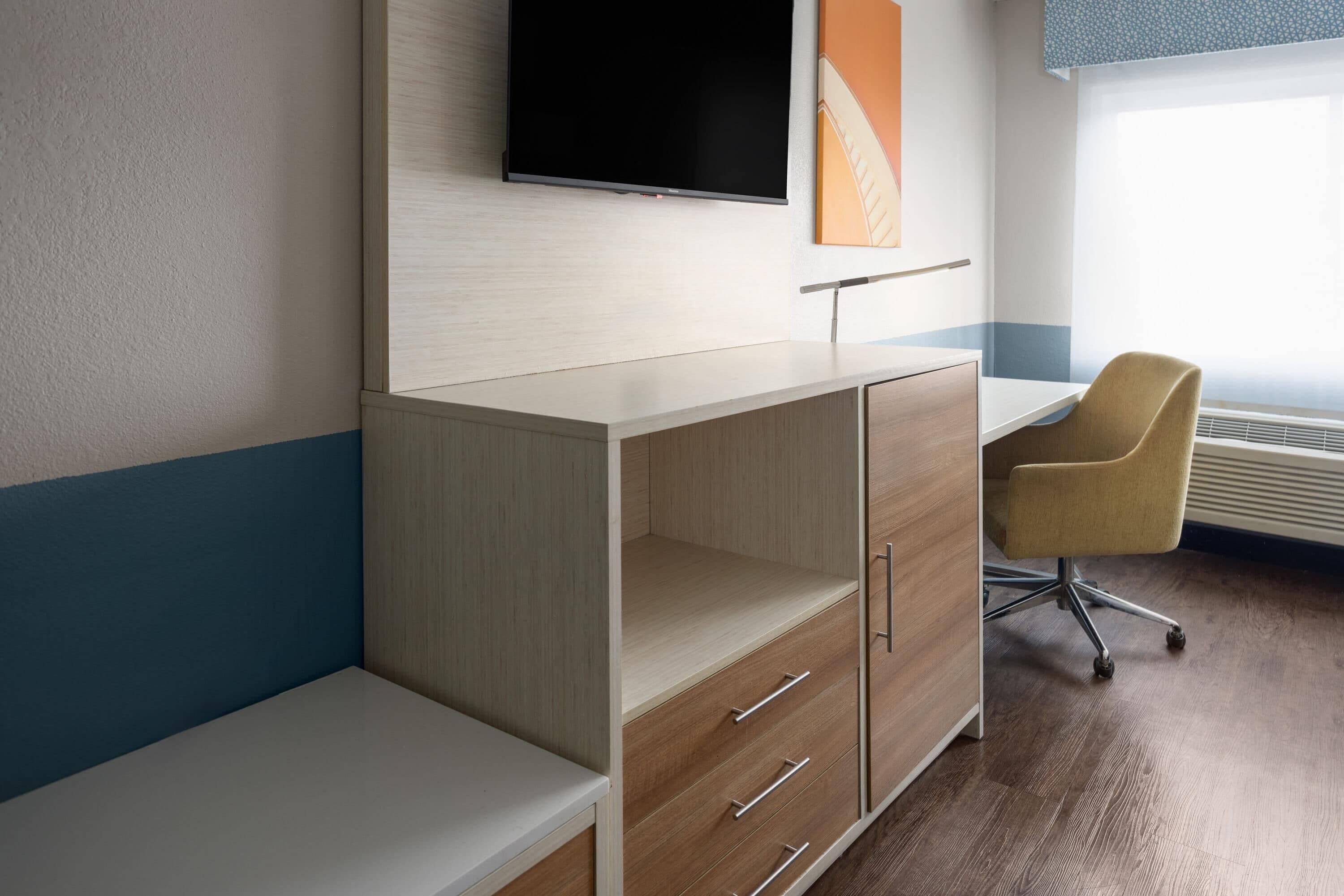 A modern hotel room with a wooden desk, a chair, a flat-screen TV, and a large window with blinds.