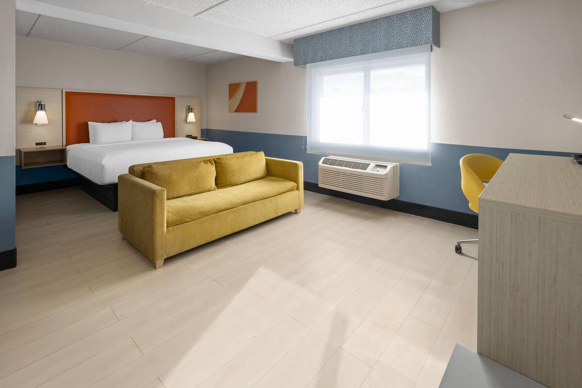 Une chambre d’hôtel avec un lit, un canapé jaune, un bureau et une fenêtre.