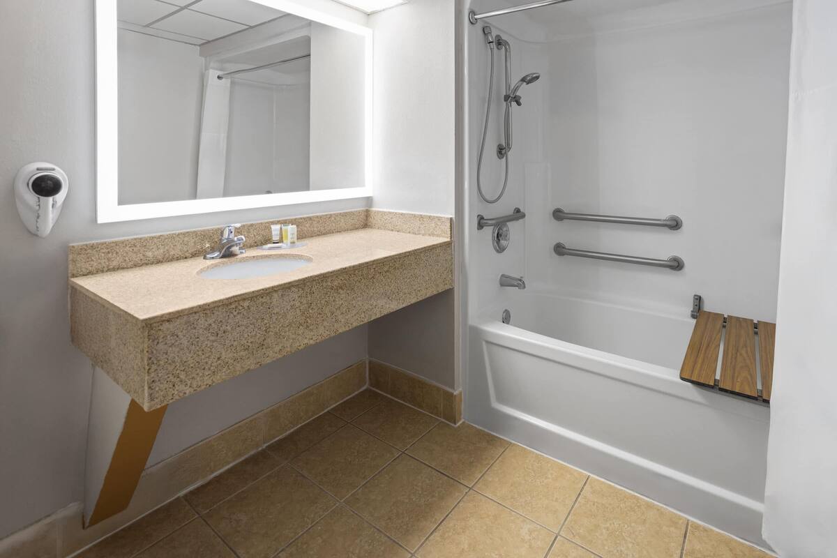 Une salle de bain avec un lavabo surmonté d’un plan de travail en granit, un grand miroir, une douche équipée de barres d’appui et une étagère en bois.