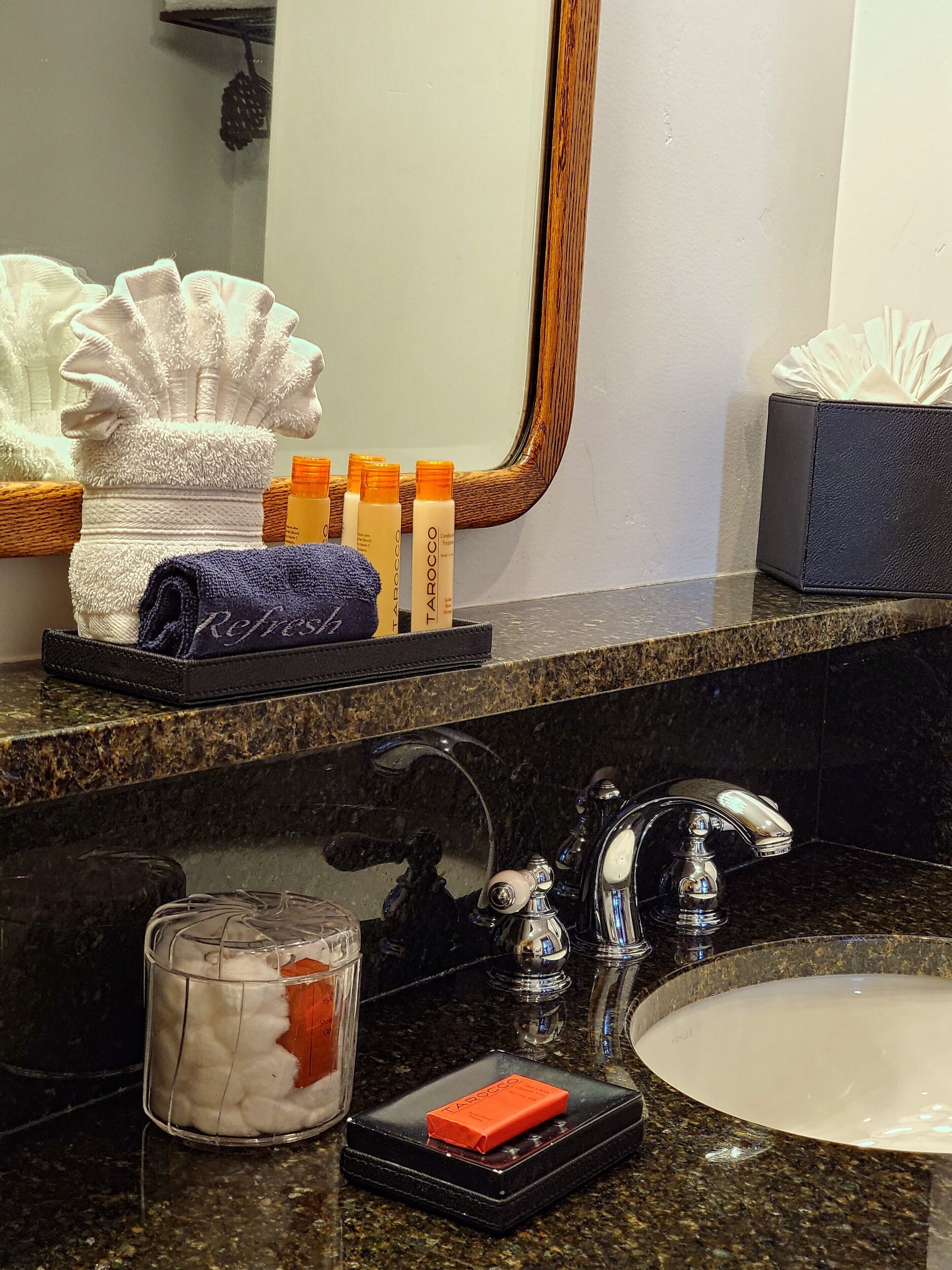 A bathroom with a marble countertop, a sink, a mirror, and toiletries including a towel, bottles, and a tissue box.