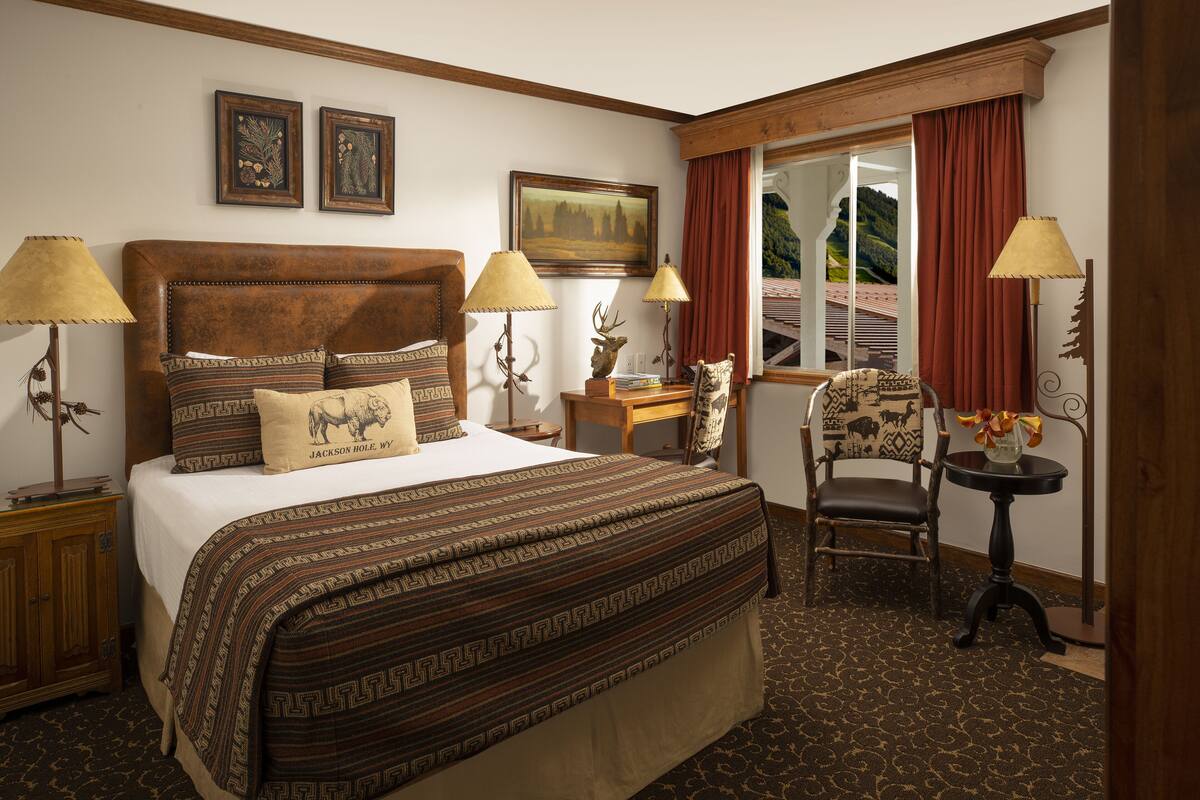 A hotel room with a large bed, two bedside lamps, a wooden headboard, a chair, a small table, and a window with curtains.