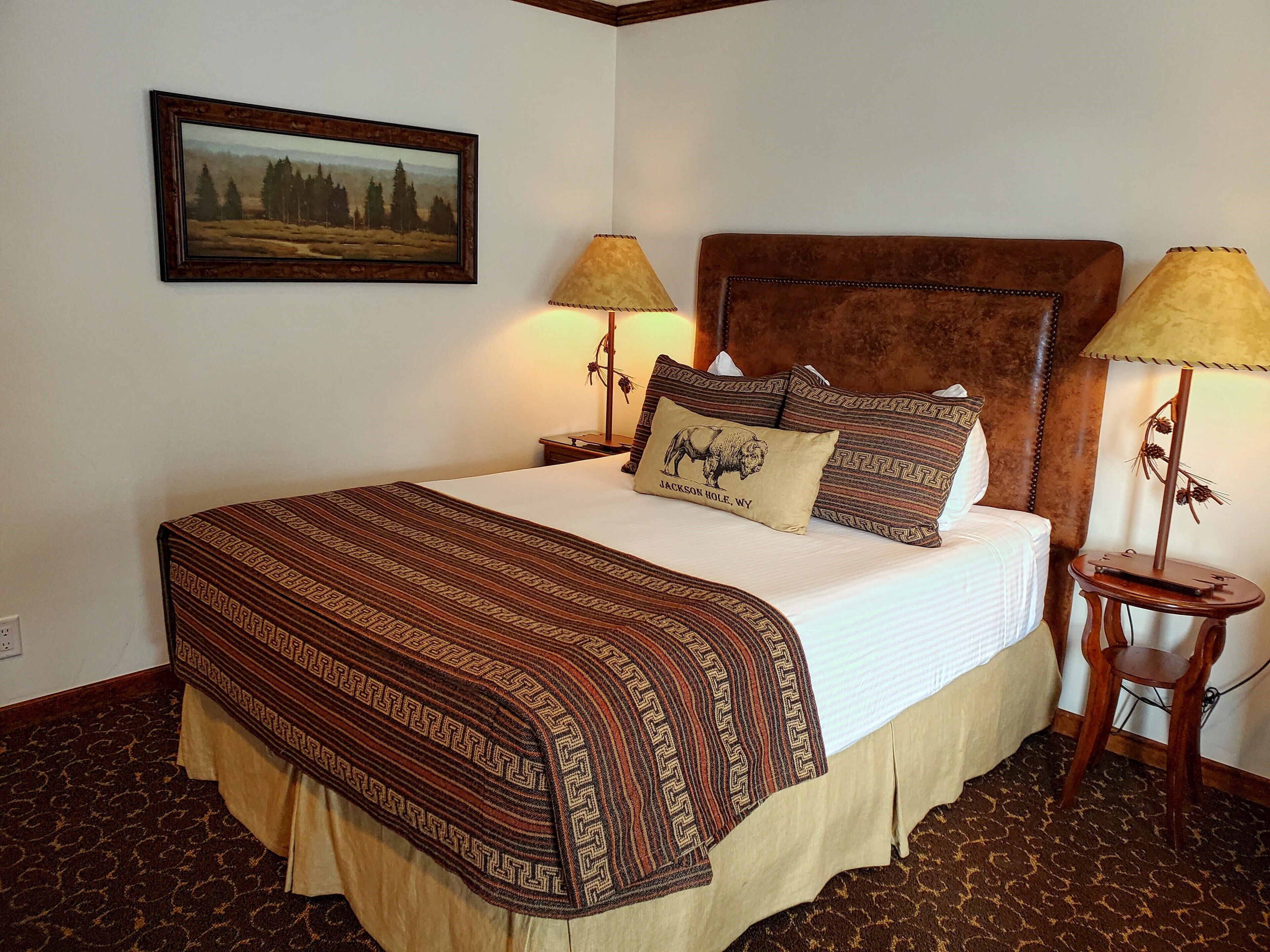 A neatly made bed with a patterned bedspread, two pillows, and a framed landscape painting on the wall.