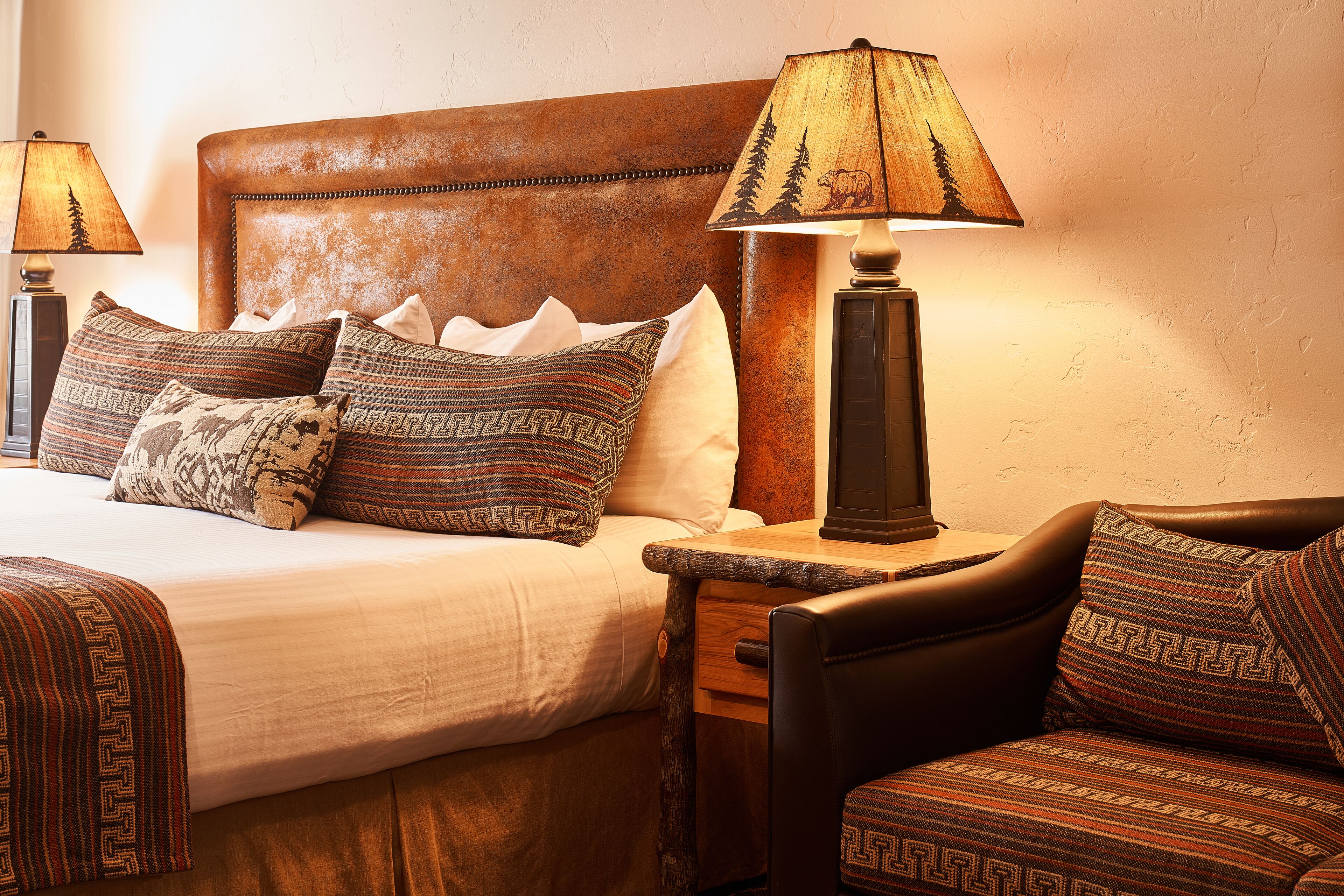A neatly made bed with patterned pillows, a wooden nightstand with a lamp, and a leather armchair.
