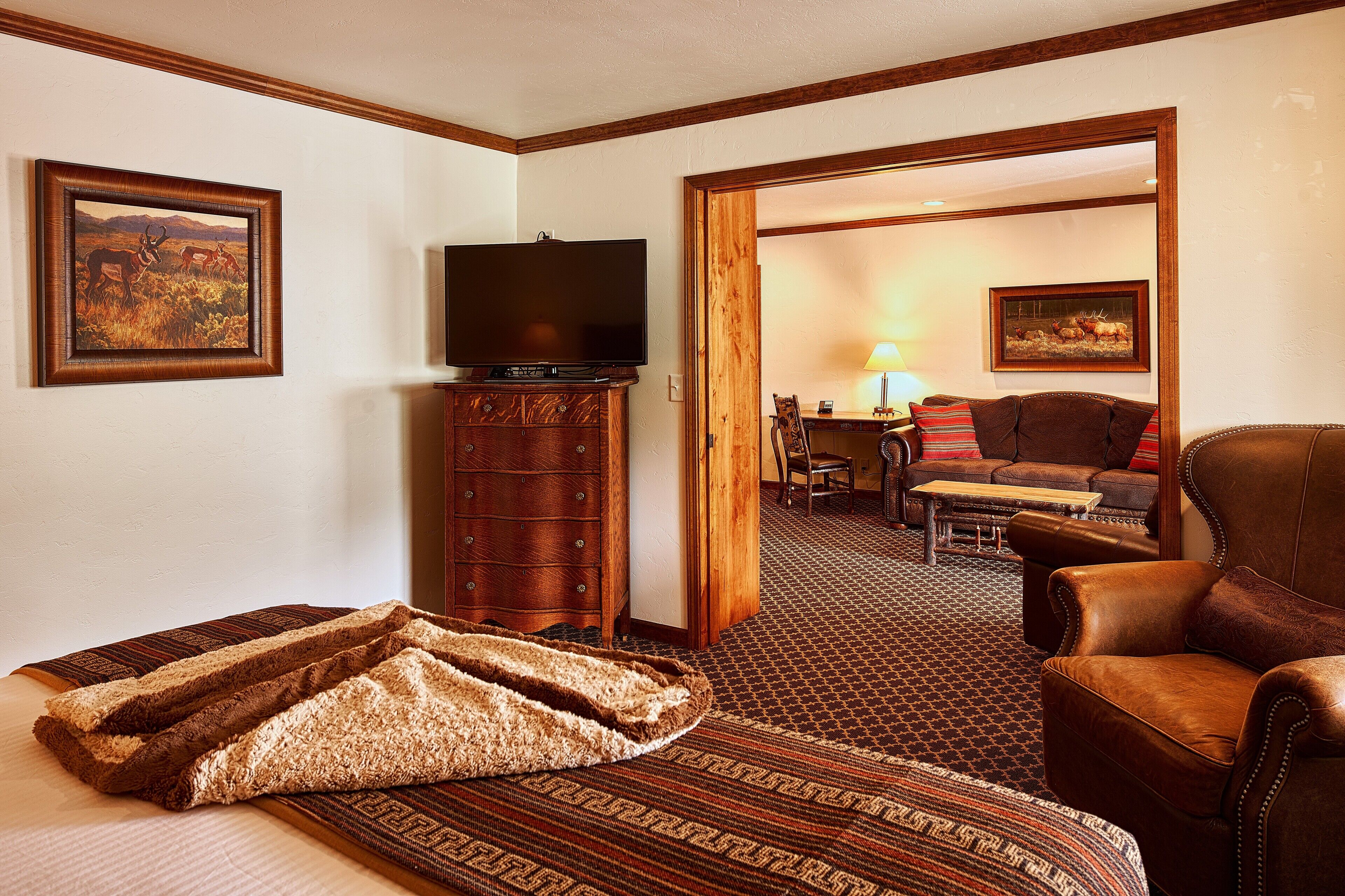 A hotel room with a bed, a television, a wooden dresser, a leather armchair, and a dining area with a table and chairs.