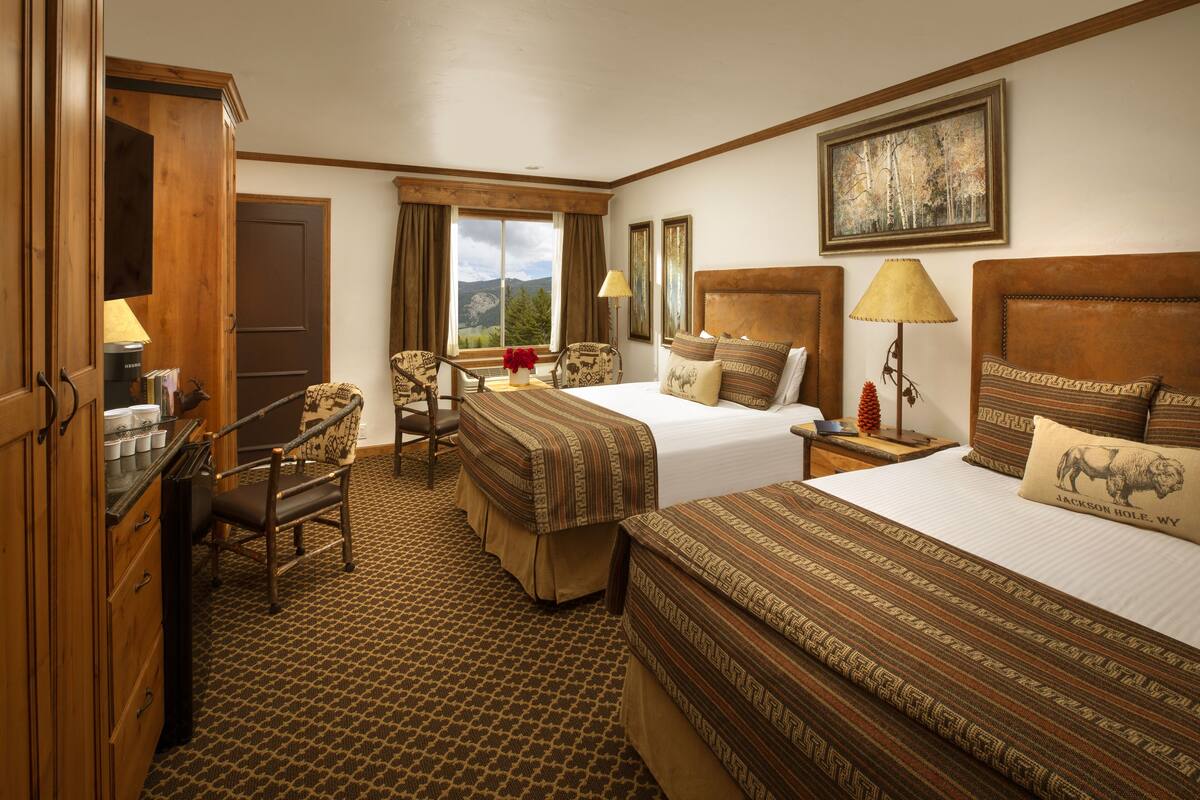 A hotel room with two beds, a desk, a chair, and a window with a mountain view.