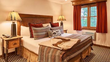 Una cama bien hecha con ropa de cama estampada, dos lámparas de noche, un teléfono en la mesita y una ventana con cortinas.