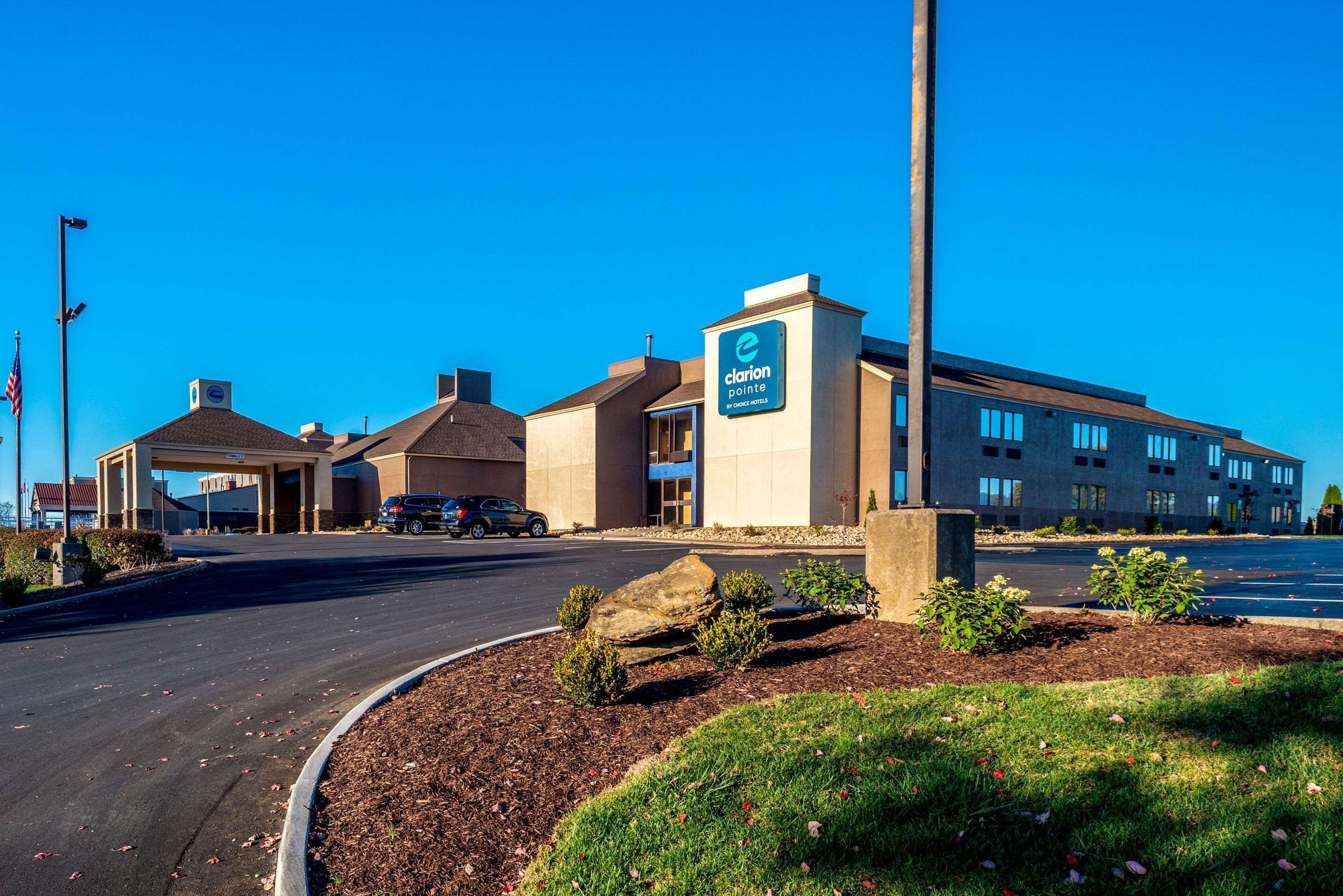A Clarion Pointe hotel with a modern design, a parking lot, and a landscaped area with small shrubs and a large rock.