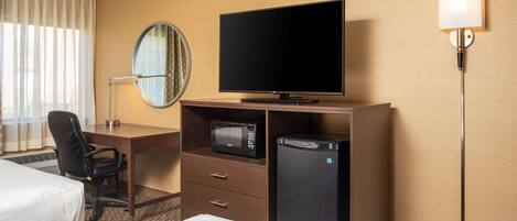 Habitación de hotel con cama, escritorio con televisor, microondas, refrigerador, silla y mesita de noche.