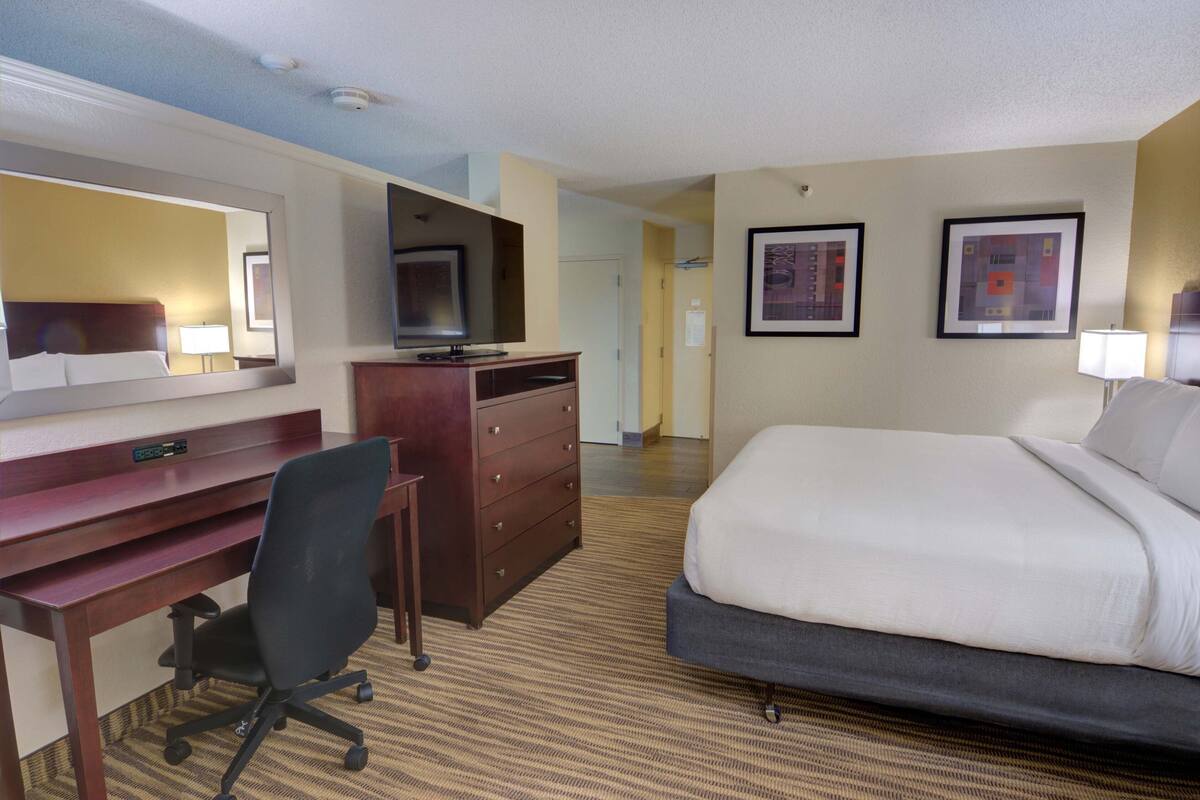 Een hotelkamer met een bed, bureau, stoel, kapstok, televisie en een kunstwerk in een lijst aan de muur.