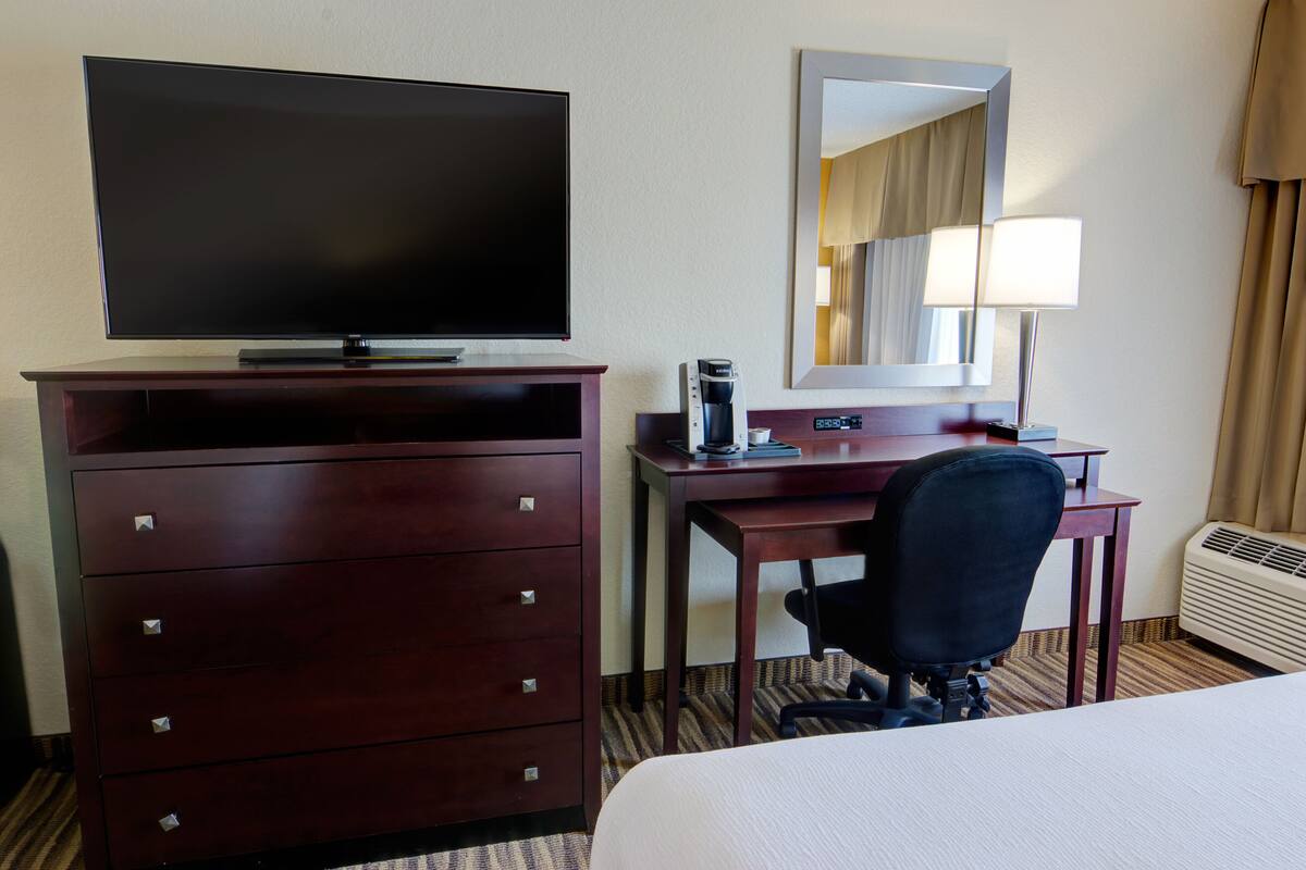 Een hotelkamer met een bed, bureau, stoel, televisie, kast, spiegel, lamp en koffiezetapparaat.
