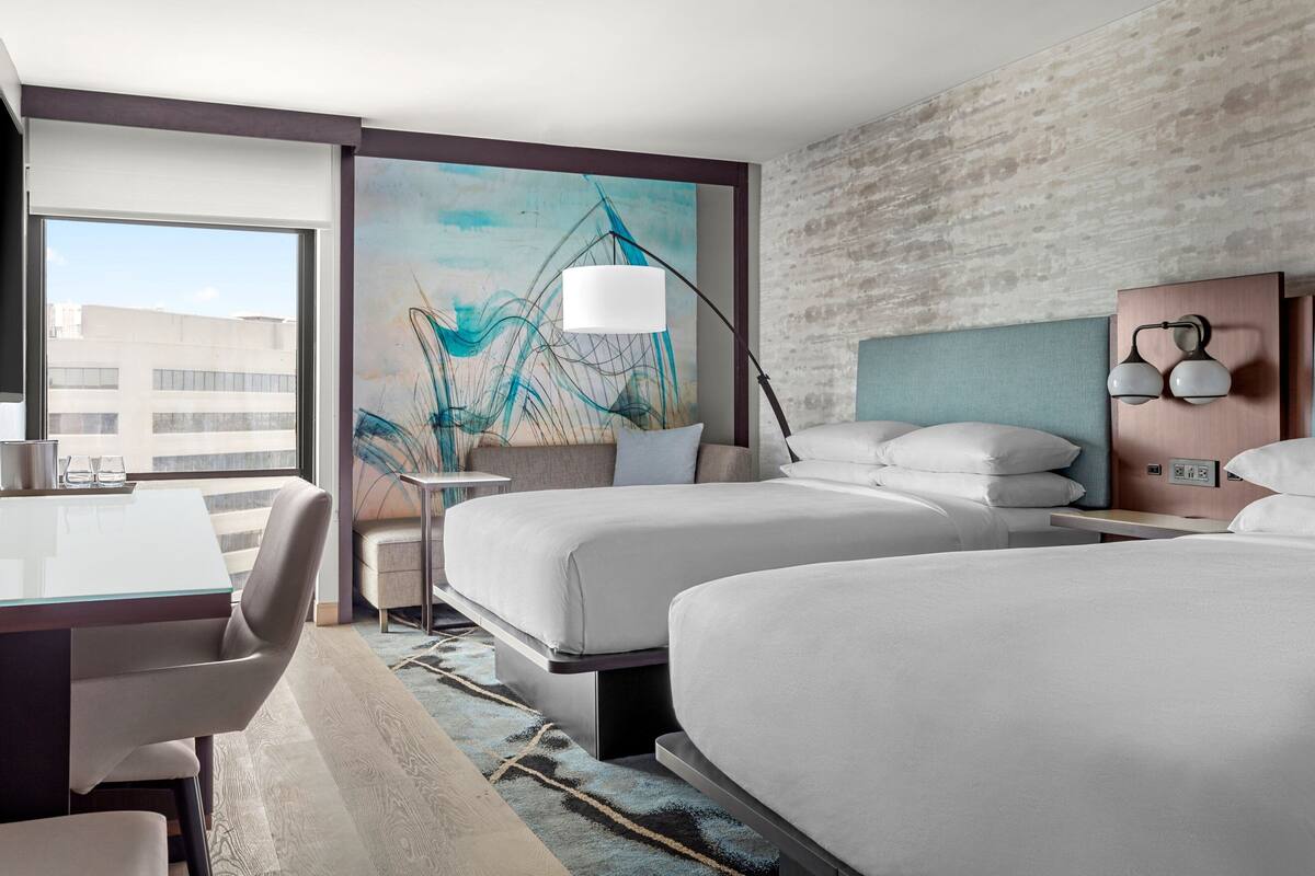 Quarto de hotel com duas camas, uma mesa, uma cadeira, um abajur e um quadro na parede.