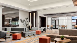 Un espacioso vestíbulo de hotel con diseño moderno, que cuenta con un amplio espacio de descanso con sofás y otomanas, una zona de bar y un ventanal con vistas a la ciudad.