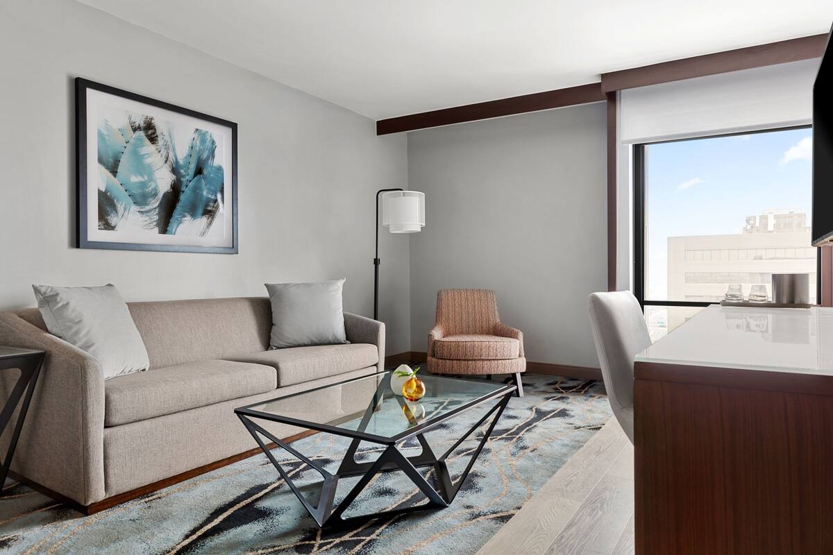 Quarto de hotel moderno com sofá, mesa de centro de vidro, cadeira, escrivaninha e uma ampla janela com vista para a cidade.
