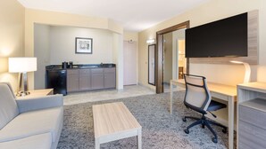 A hotel room with a sofa, a small coffee table, a desk with a chair, a flat-screen TV mounted on the wall, and a kitchenette in the background.