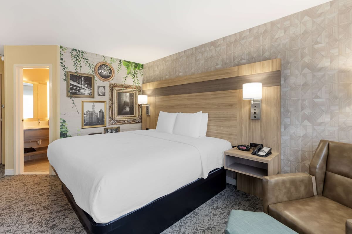 A hotel room with a large bed, a wooden headboard, a nightstand with a lamp, a telephone, and a seating area with a chair and ottoman.
