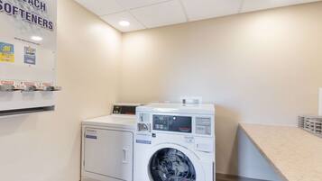 A laundry room with a front-loading washer and dryer, a wall-mounted detergent dispenser, and a sign advertising bleach softeners.