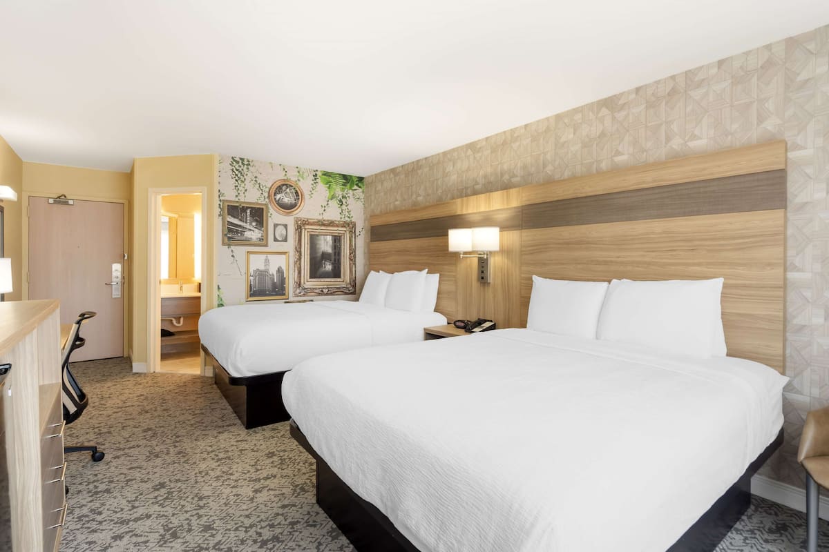 A hotel room with two beds, a desk, a chair, a lamp, and a wall with framed pictures and a plant.