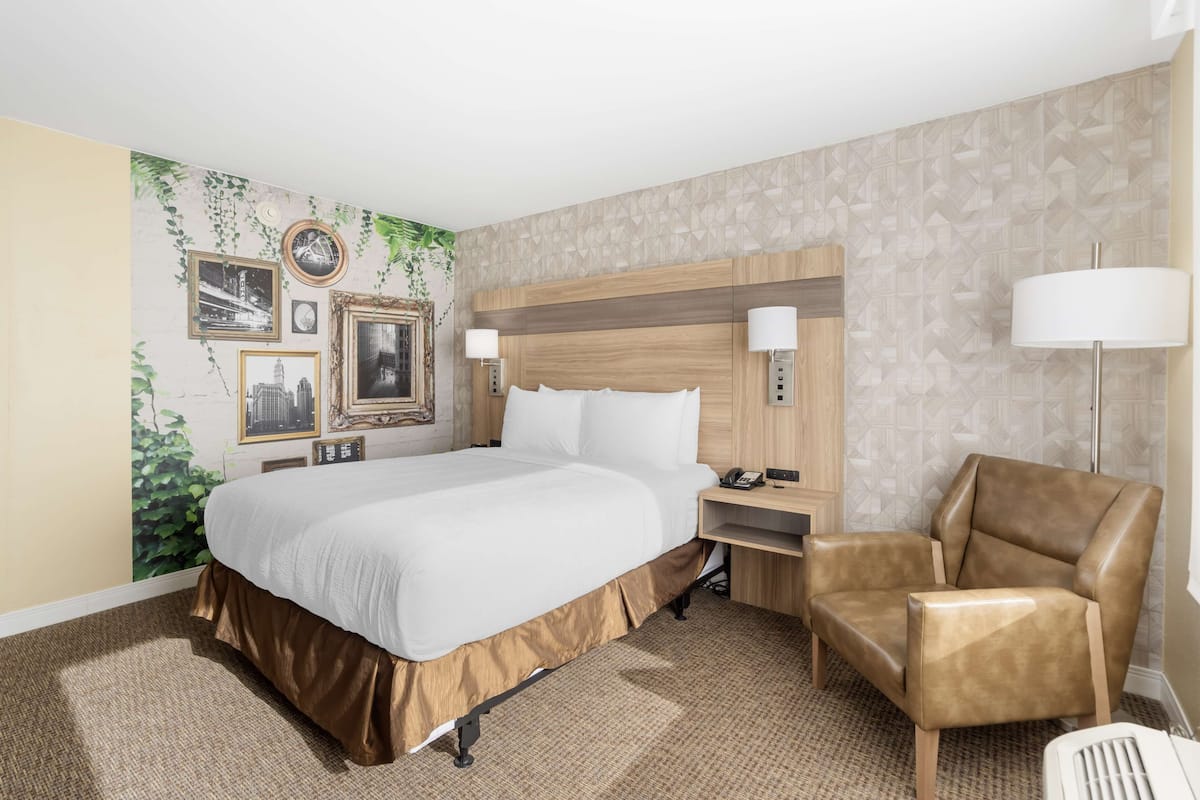 A hotel room with a large bed, a wooden headboard, a brown leather chair, and a wall with framed pictures and a clock.