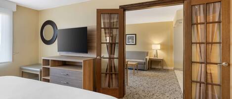 A hotel room with a bed, a desk with a television, a chair, a lamp, and a door leading to another room.