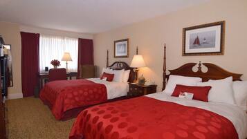 A hotel room with two beds, each with red bedding and white pillows, a wooden headboard, a bedside table with a lamp, and a framed picture on the wall.
