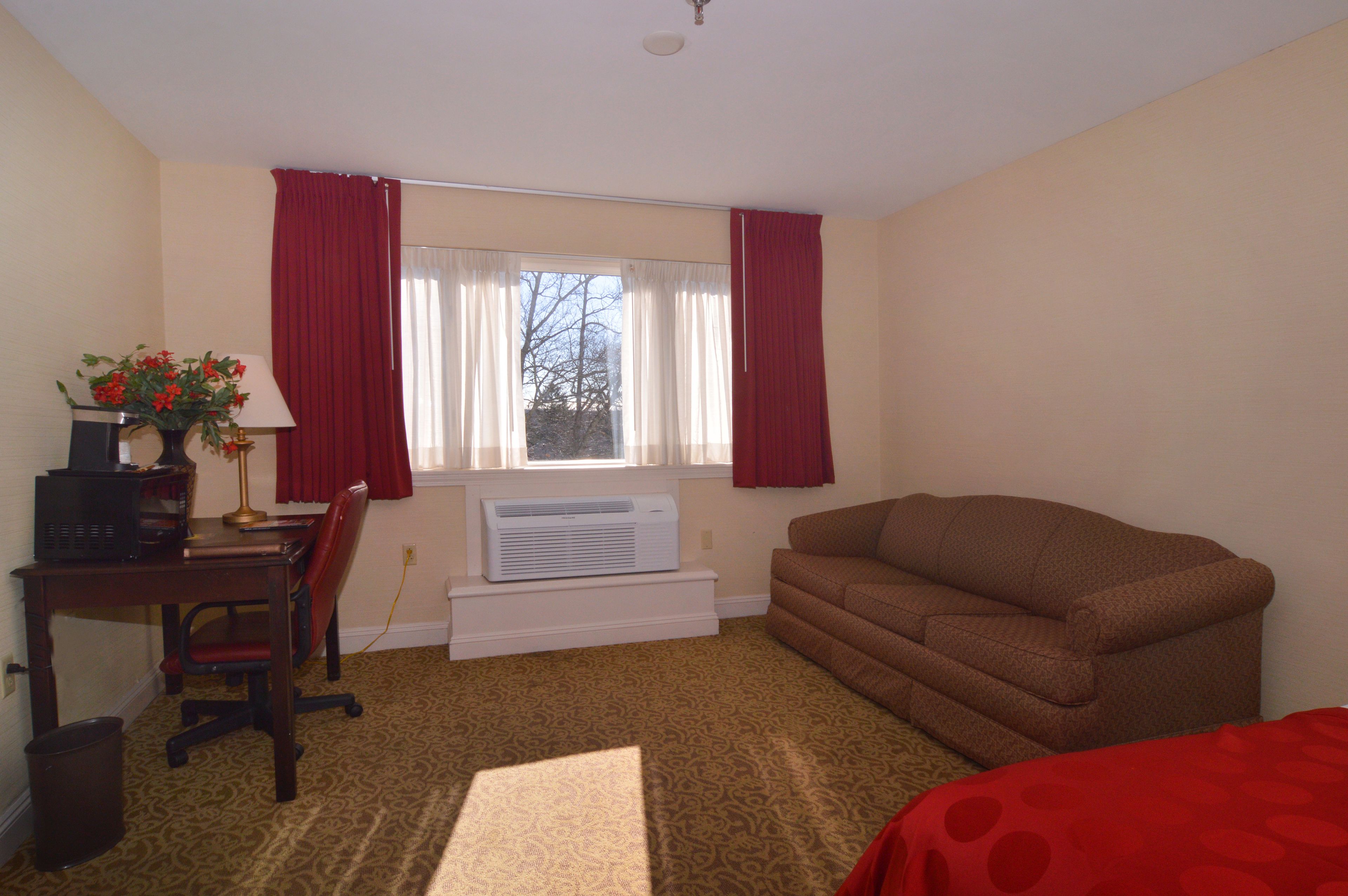Una habitación con un sofá marrón, un escritorio con computadora y lámpara, una ventana con cortinas rojas y un radiador.