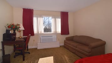 A room with a brown couch, a desk with a computer and a lamp, a window with red curtains, and a radiator.