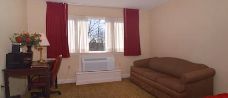 Una habitación con un sofá marrón, un escritorio con computadora y lámpara, una ventana con cortinas rojas y un radiador.
