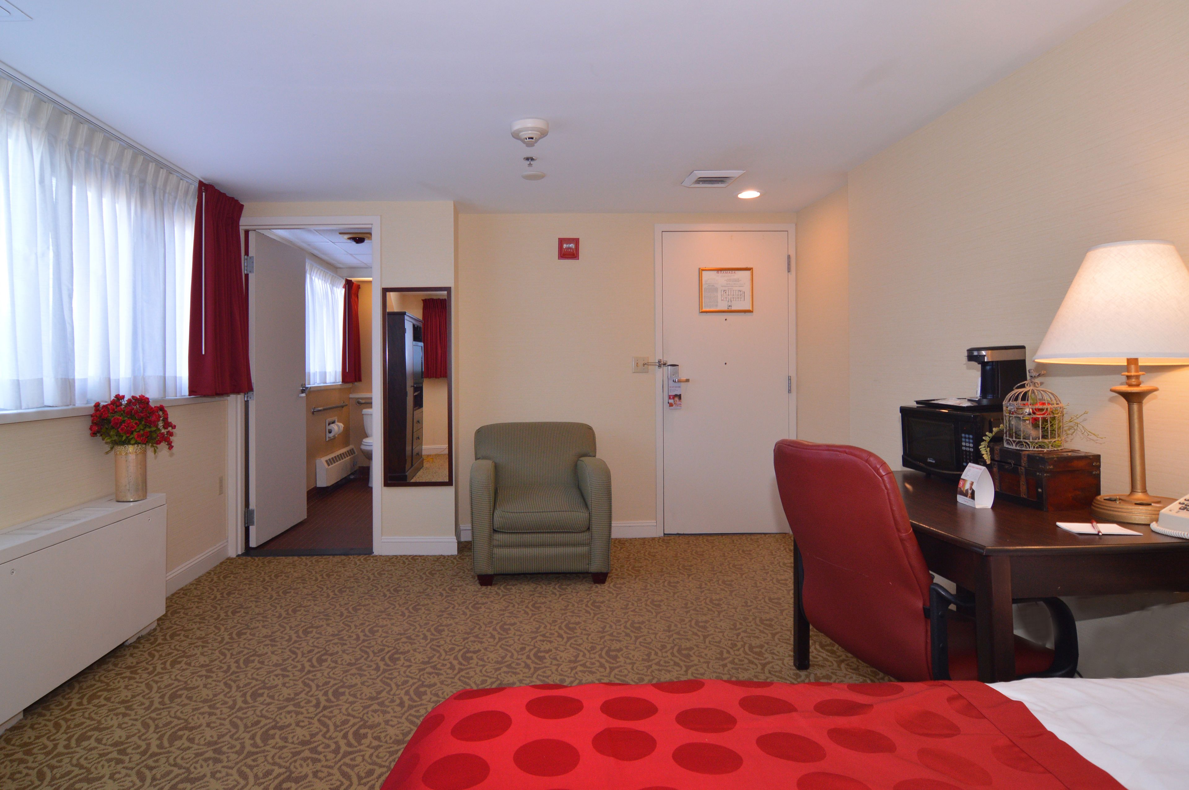 A hotel room with a bed, a desk with a lamp, a chair, a small table with a decorative item, a red polka dot bedspread, a vase with flowers, and a door leading to a bathroom.