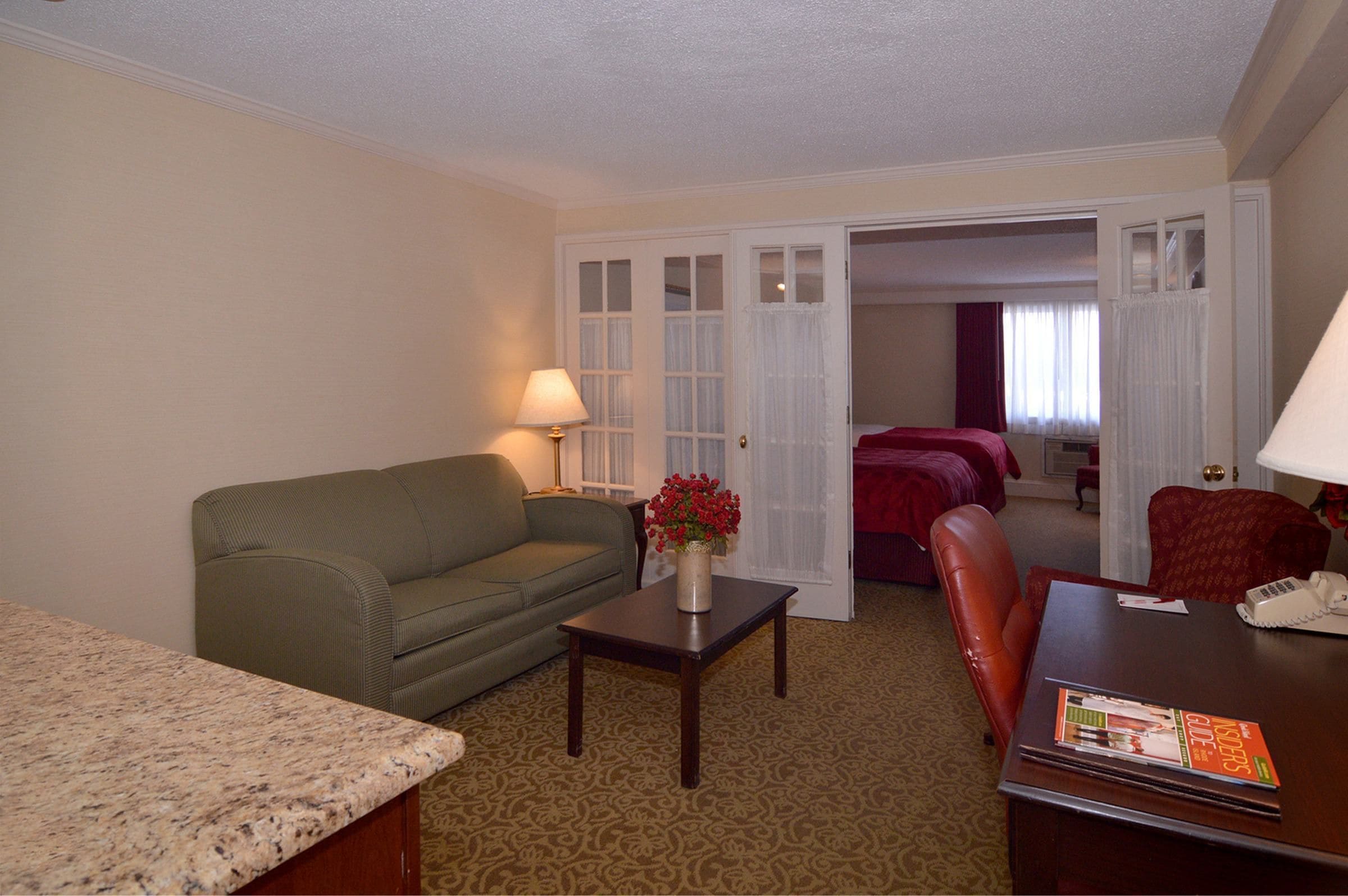 A hotel room with a green sofa, a small table with a vase of flowers, a wooden desk with a telephone and a magazine, and a bed with a red cover.