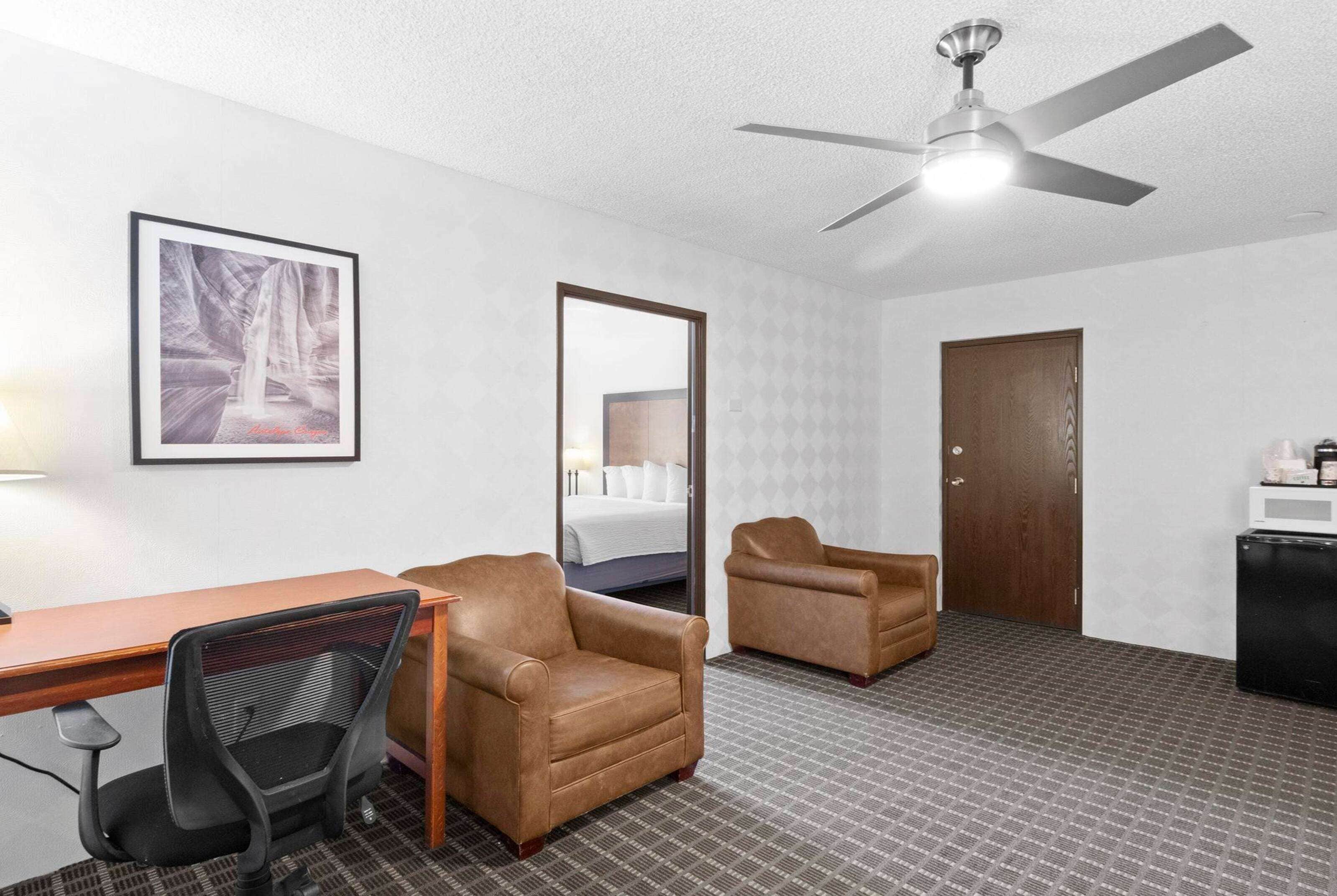 A hotel room with a desk, two leather armchairs, a black office chair, a ceiling fan, a framed picture on the wall, a door, and a small kitchenette.