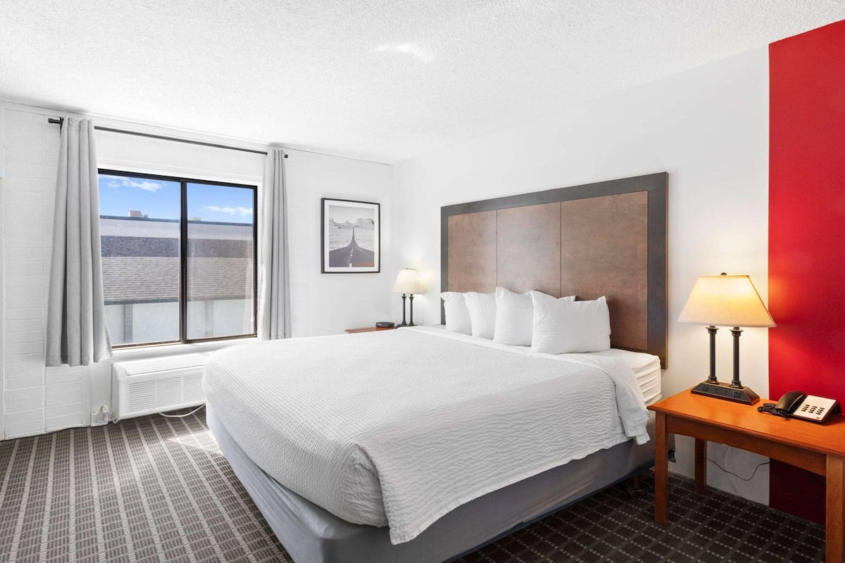 A hotel room with a large bed, two bedside tables with lamps, a window with curtains, a framed picture on the wall, and a red accent wall.