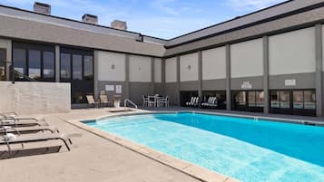 Una piscina con lettini da relax e un tavolo, circondata da un edificio con ampie finestre e facciata in mattoni.