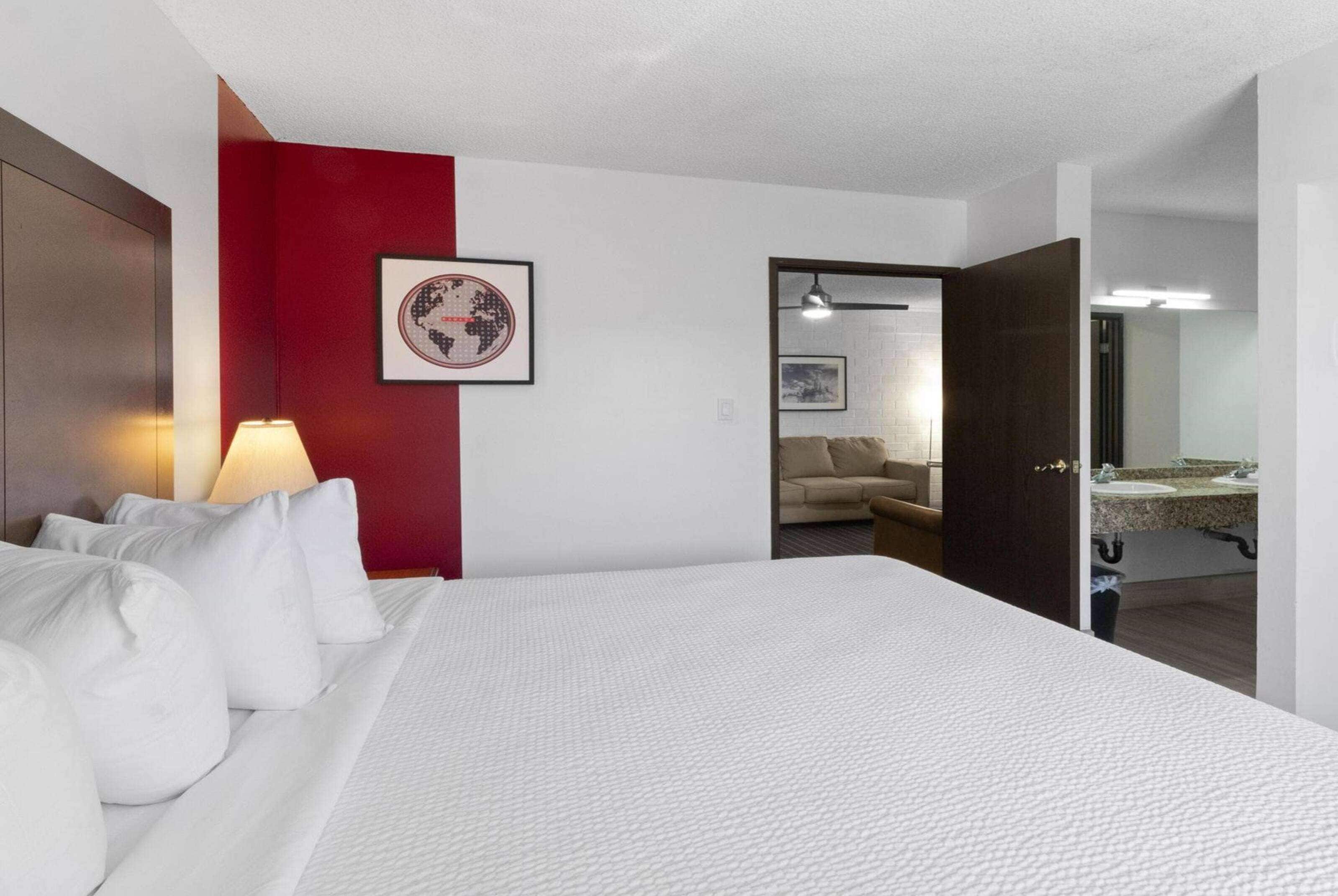 A hotel room with a large bed, a red accent wall, a framed world map, a sofa, and a bathroom visible through an open door.