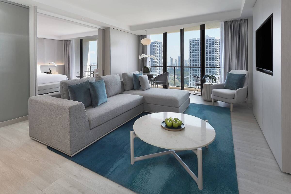 A modern hotel room with a grey sectional sofa, a round coffee table, and a balcony with a view of the city.