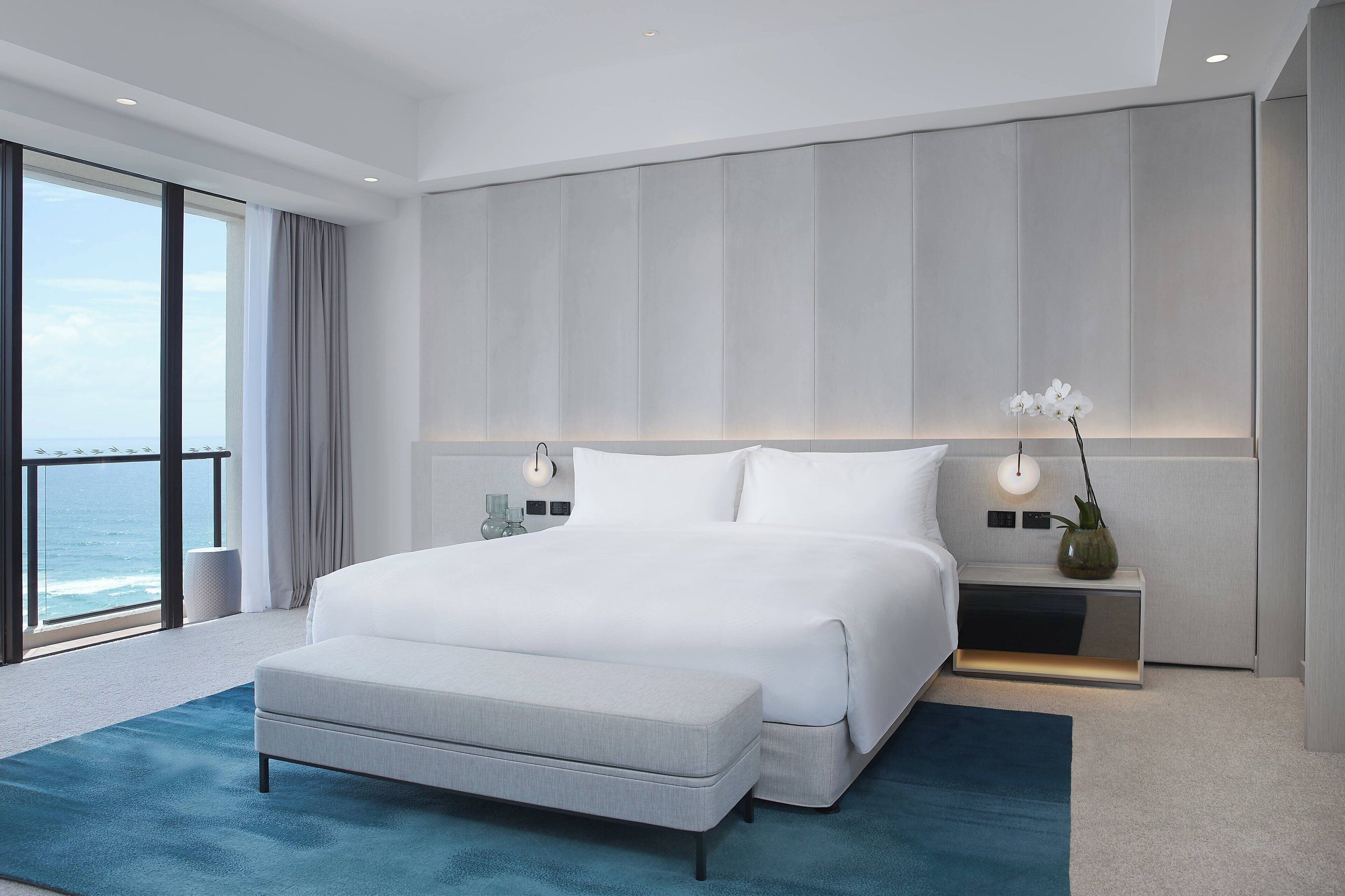 Une chambre d’hôtel moderne avec un grand lit, une table de chevet avec une lampe, un vase de fleurs et une vue sur l’océan.
