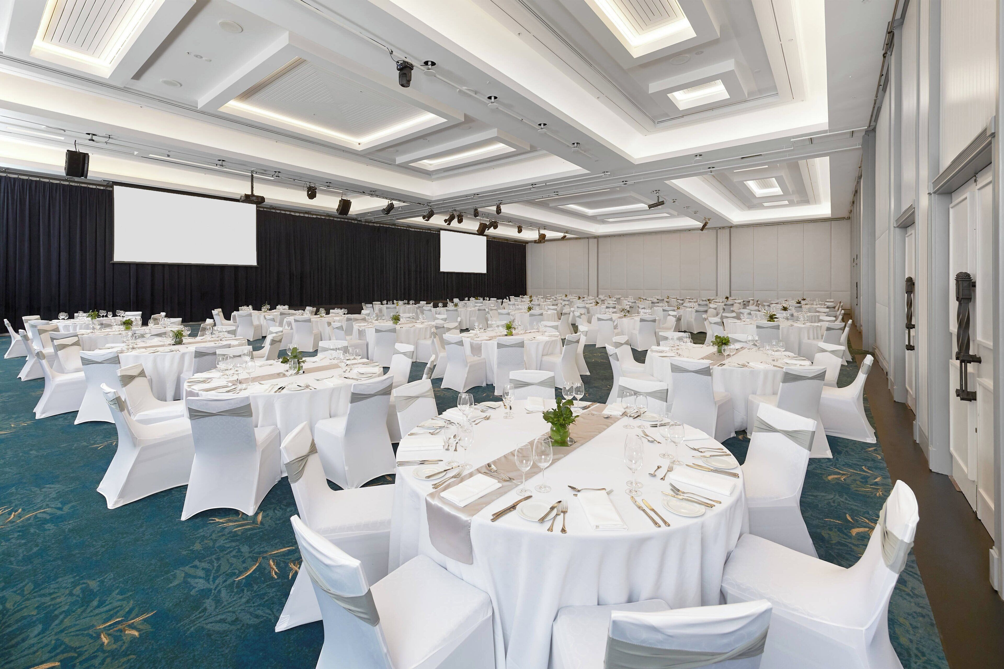 Un amplio salón de conferencias con filas de mesas redondas, sillas blancas y un escenario con una pantalla.