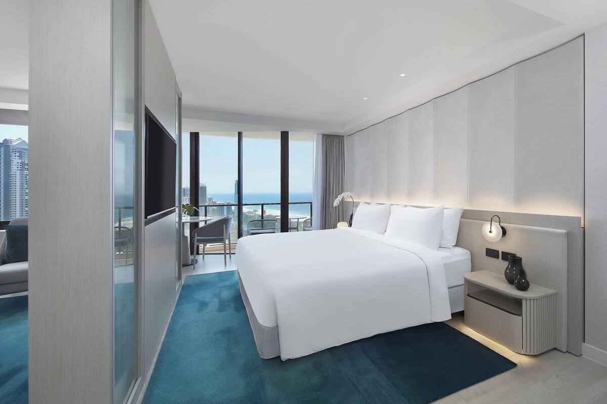 A modern hotel room with a large bed, a bedside table with a lamp, a TV, and a balcony with a city view.
